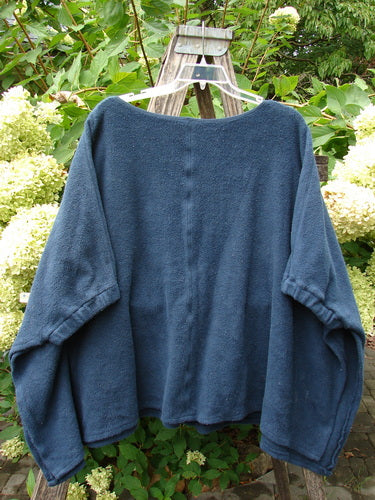 Barclay Celtic Moss Patched Poncho Single Six Daisy Ink Blue OSFA displayed on a white hanger, showcasing its open sides and gathered lower sleeves, set against a garden backdrop with green foliage and hydrangeas.