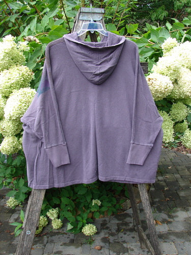 Barclay NWT Cotton Hemp Folklore Hoodie Morning Dove Aura OSFA, a loose-fitting, long-sleeved hoodie with a kangaroo pocket, displayed on a wooden frame amidst green foliage and white hydrangeas outdoors.