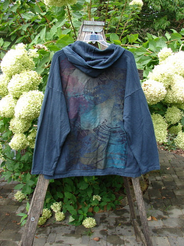 Barclay Cotton Hemp Hip To Be Square Hoodie Rear Botanicals Black OSFA displayed on a stepladder, showcasing its multicolored graphic print on the back amidst a lush garden setting.