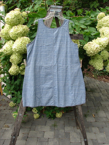 Barclay NWT Light Weight Chambray Shift Stripe Denim Light Size 0 hangs on a wooden frame, showcasing its sleeveless A-line design, scoop neckline, and unique varying hem against a natural backdrop.