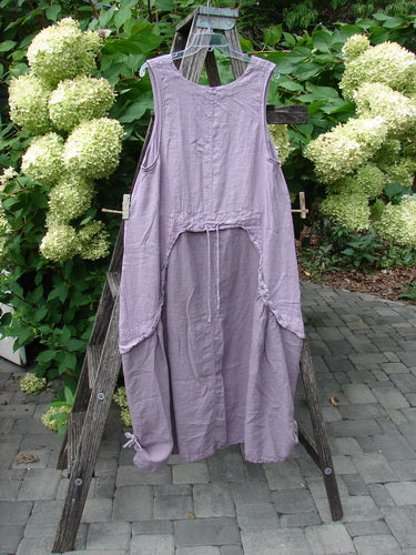 Barclay NWT Linen Explorer Jumper Tiny Botanical Sea Fog Size 0: A loose lavender linen smock with adjustable ties, side pockets, and scoop neckline, displayed on a wooden A-frame amidst lush hydrangeas.