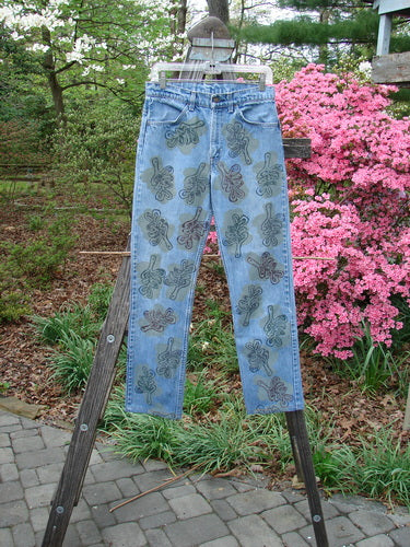 1995 Levi Strauss & Co Denim Blue Jean Leaves Stone Wash Size 1, featuring abstract floral motifs, hangs from a wooden ladder outdoors, showcasing its unique vintage style amidst lush green foliage and pink blossoms.