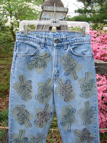 1995 Levi Strauss & Co Denim Blue Jean Leaves Stone Wash Size 1, featuring an abstract leaf motif in greenish-brown, displayed on a hanger amid a garden setting with pink flowering shrubs.