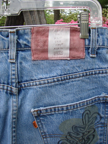 1995 Levi Strauss & Co Denim Blue Jean Leaves Stone Wash Size 1, featuring a pinkish label, orange tab, and decorative leaf graphic, showcased hanging outdoors against a floral, green backdrop.