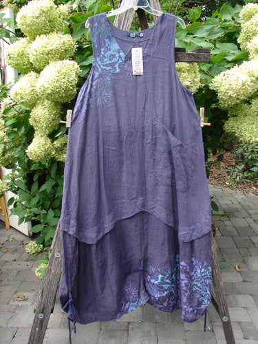Barclay NWT Linen Explorer Jumper in Purple River Mist hangs on a trellis, showcasing its scoop neckline, tiered hemline, and botanical blossom accents. The dress features side pockets and retains its store tag.