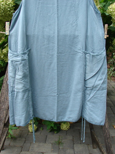 Barclay NWT Lyocell Joyous Jumper Unpainted Sea Green Size 0 displayed on a wooden rack, featuring two large patch pockets, side ties, and a knee-length A-line shape, secured with clothespins.