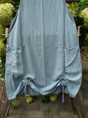 Barclay NWT Lyocell Joyous Jumper Unpainted Sea Green Size 0, featuring a loose fit, sleeveless design with patch pockets and drawstring hem, displayed against a backdrop of green foliage and flowers.