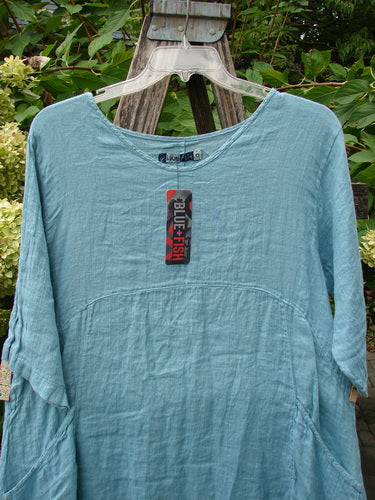 Barclay NWT Linen Zen Pocket Tunic Dress Unpainted Turquoise Size 0, featuring three-quarter sleeves, A-line shape, dual pockets, and varying hemline, displayed on a hanger against lush greenery.