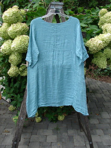 Barclay NWT Linen Zen Pocket Tunic Dress in turquoise, size 0, displayed outdoors on a wooden ladder. Features include a rounded neckline, three-quarter sleeves, dual pockets, and varying hemline.