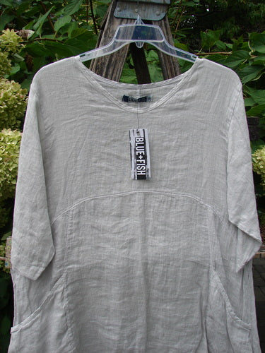 Barclay NWT Linen Zen Pocket Tunic Unpainted Dove Size 0 hangs on a clear hanger. Features include three-quarter sleeves, dual pockets, and an A-line shape, showcasing Blue Fish's creative, vintage style.
