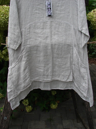 Barclay NWT Linen Zen Pocket Tunic Unpainted Dove Size 0 on an outdoor wooden rack, featuring ¾-length sleeves, asymmetrical hem, rounded yoke seam, and front patch pockets with a BLUE FISH tag.
