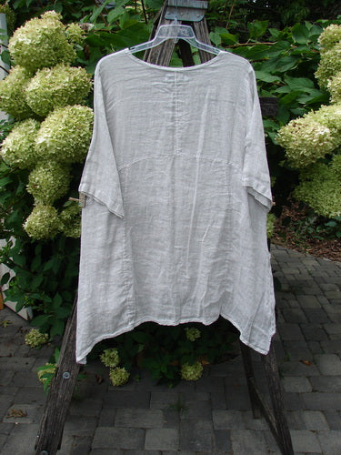 Barclay NWT Linen Zen Pocket Tunic Unpainted Dove Size 0 hanging on a wooden ladder, showcasing its loose fit, three-quarter sleeves, and A-line shape amidst lush hydrangeas in an outdoor setting.