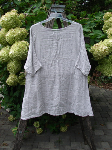 Barclay NWT Linen Zen Pocket Tunic Unpainted Pearl Smoke Size 0, displayed on a wooden A-frame, features a loose fit, three-quarter sleeves, dual pockets, and a varying hemline.