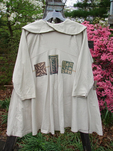 1994 Wanderer's Jacket Mindful Mosaic Mist Altered Size 1 displayed on a swing, showcasing its wide shawl collar, sectional side inserts, and mosaic theme paint, amidst a garden setting with pink flowers.