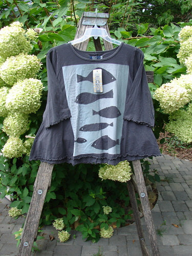 Barclay NWT Three Quarter Sleeved Crop Cafe Tee Many Fish Black Sand Size 0, featuring a loose-fit, dark gray top with fish silhouettes, displayed on a ladder outdoors amidst greenery.
