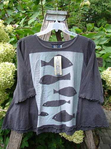 Barclay NWT Three Quarter Sleeved Crop Cafe Tee hangs on a wooden garden ladder, showcasing bell sleeves, fish silhouettes on the front, and a tag amid lush greenery and white hydrangeas.