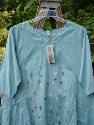 Barclay NWT Batiste Cross Over Urchin Pocket Tunic Tiny Tulip Turquoise Size 0, featuring ¾-length sleeves, unique sectional panels, two side pockets, and vibrant floral embroidery, hanging on a clear hanger outdoors.