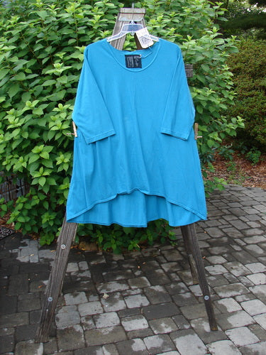 Barclay NWT High Low Top Unpainted Pacific Aqua Size 2 displayed on a wooden rack, showcasing its A-line shape, rounded banded bottom, and varying hemline with three-quarter sleeves.
