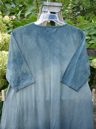 Barclay NWT Wander Tunic Tee in Shadow Blossom Mottled Grey Green hangs on a plastic hanger outdoors, showcasing its mid-length, loose fit, elbow-length sleeves, and organic cotton fabric.