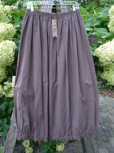 Barclay NWT Batiste Airy Lawn Skirt Unpainted Brown Sparrow Size 0, featuring an elastic waistband and hem, hangs on a clear hanger, showcasing its lightweight, gathered design against a garden backdrop.