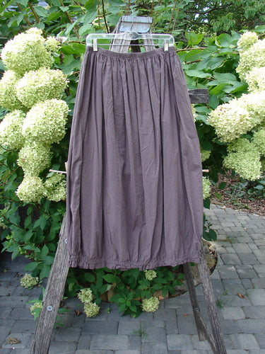 Barclay NWT Batiste Airy Lawn Skirt Unpainted Brown Sparrow Size 0, displayed from a plastic hanger on an outdoor ladder, amidst lush greenery and hydrangeas, showcasing its loose, airy barrel shape.