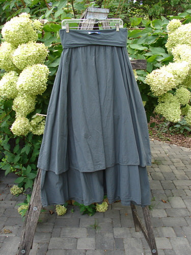 Barclay NWT Batiste Fold Over Stardust Tier Skirt in Atlas Grey size 0, displayed on a wooden A-frame, features a flowing, tiered design with a full-length silhouette amidst a garden backdrop.