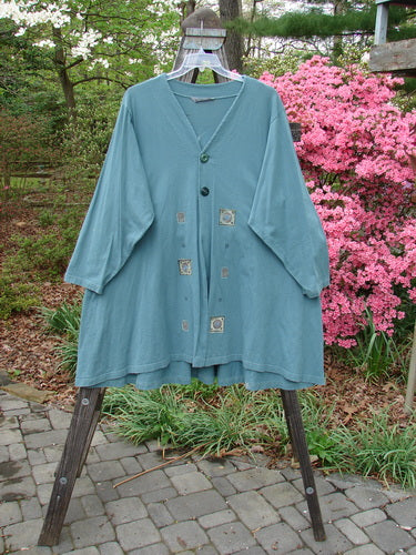 1997 Mask Jacket Quad Celtic Knot Cypress Size 2 on a wooden rack, featuring a bell shape with two button closure, two deep side pockets, and a rounded swing hemline.