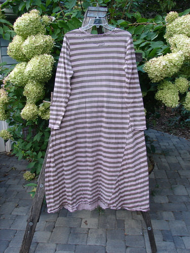 The Magnolia Pearl Cotton Dylan Dress Hand Mended Lavender Stripe OSFA hangs elegantly on a wooden ladder, showcasing its long sleeves, horizontal stripes, rounded neckline, and distinctive hand-mended details.