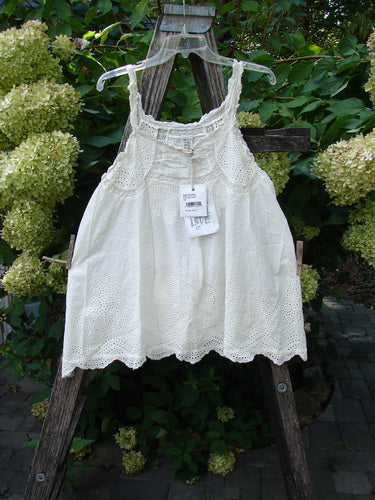 Magnolia Pearl NWT Embroidered Eyelet Cotton Clementine Tank True OSFA, featuring intricate eyelet detailing around the neckline and hem, hangs on a rustic wooden trellis, highlighting its pintucked front and lace-adorned straps.