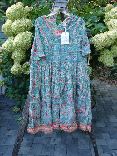 Magnolia Pearl NWT European Cotton Block Print Taigi Dress Demi OSFA displayed on a hanger, showcasing its floral pattern, pintucks, pleats, and unique ric rack accents, set against a serene garden backdrop.