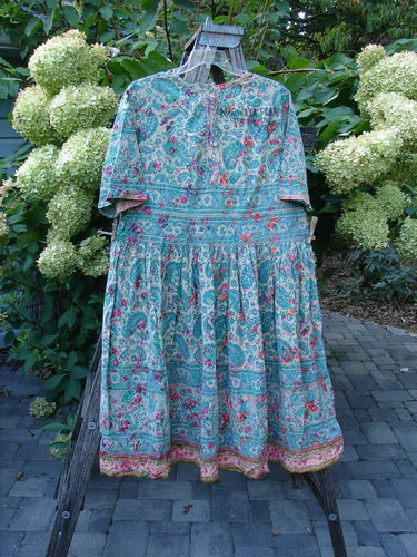 Magnolia Pearl NWT European Cotton Block Print Taigi Dress Demi OSFA, featuring elbow-length sleeves, a full gathered skirt, and detailed pintucks, hangs on a wooden stand, showcasing its vibrant floral and paisley pattern.