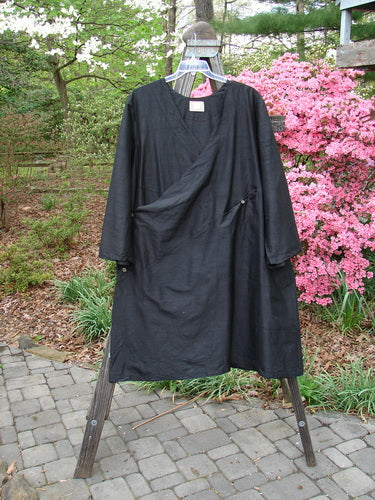 2000 Shaunting Silk Slant Wrap Jacket Unpainted Deep Black Size 2, displayed on a wooden frame, highlighting its loose fit, wider sleeves, and slightly shimmery texture against a lush outdoor backdrop.