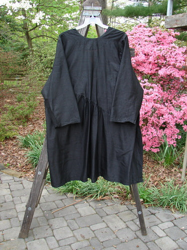 2000 Shaunting Silk Slant Wrap Jacket Unpainted Deep Black Size 2 displayed on a wooden A-frame outdoors, highlighting its loose fit, long sleeves, and billowy Shantung silk texture.