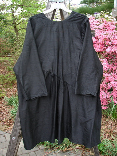 2000 Shaunting Silk Slant Wrap Jacket Unpainted Deep Black Size 2 hangs outdoors, showcasing its billowy silk, wide rounded sleeves, and front slit pockets against a lush garden backdrop with pink blooms.