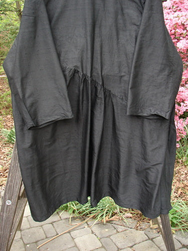 The 2000 Shaunting Silk Slant Wrap Jacket, in unpainted deep black, is displayed on an outdoor wooden ladder, showcasing its loose fit, diagonal seam, and slightly shimmery, textured fabric.