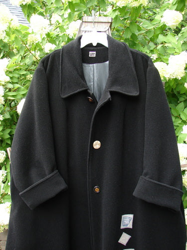 Black 1999 Patched Woven Overcoat, Size 2, displayed on a swinger. Features oversized buttons, deep pockets, and hand blacked patches, showcasing its vintage-inspired, A-line design.