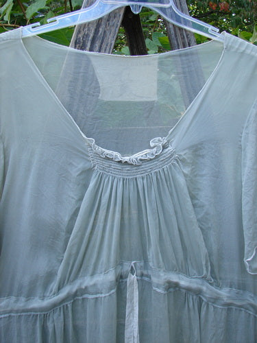 Magnolia Pearl Silk Voile Smocked Tie Back Dress in Lightest Sage, featuring a gathered bust and neckline with delicate ruffle trim, hangs on a wooden trellis, showcasing its semi-sheer, flowing design.
