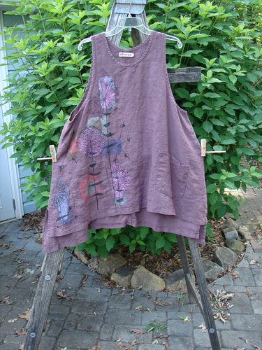 Barclay Linen Two Pocket Shirttail Pinafore Starburst Plum Wine Size 1, featuring an A-line flair and double drop exterior pockets, hangs gracefully on a wooden rack, showcasing its flowy, artistic design.