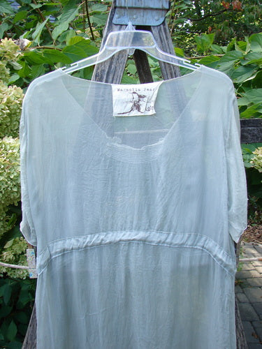 Magnolia Pearl Silk Voile Smocked Tie Back Dress Lightest Sage OSFA, semi-sheer and airy, hangs elegantly on a clear hanger, showcasing its scooped neckline, three-quarter sleeves, and delicate vertical gathers.