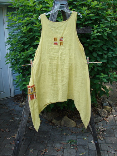 Barclay Linen Single Side Pocket Pinafore Geos Sunshine Size 1 displayed on a wooden stand, highlighting its A-line flair, deeper scoop neckline, and unique single drop side pocket with a geometric pattern.