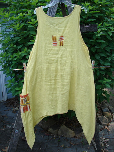 Barclay Linen Single Side Pocket Pinafore Geos Sunshine Size 1, featuring a sweeping A-line flair, deeper scoop neckline, and a unique exterior painted pocket in the Geos theme, displayed on a clothesline.