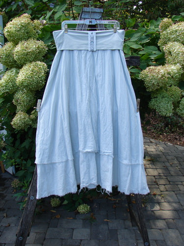 Barclay NWT Batiste Fold Over Raglan Silhouette Skirt Unpainted Dew Size 1 displayed on a hanger, showcasing its pale-blue, tiered design with frayed edges, against a lush garden backdrop.