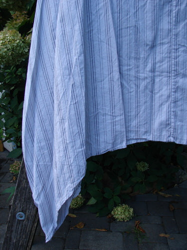 Barclay NWT Percale Hado Shift Stripe Smoke Pearl Size 1 drapes elegantly from a wooden rail, showcasing its light-blue pinstriped cotton percale fabric against a backdrop of leafy shrubs.