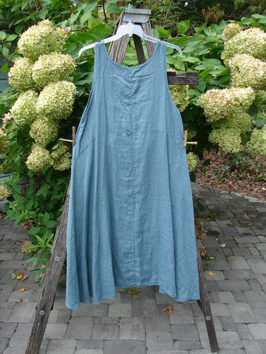 Barclay NWT Linen Tulip Midlayer Overpiece Unpainted La Mer Size 0, a sleeveless, loose-fit dress with a layered crossover front, hangs on a wooden A-frame, showcasing its sweeping A-line design.