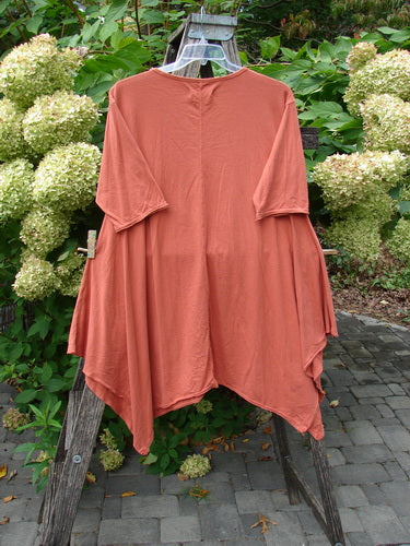 Barclay NWT Origami Tunic Top Berrie Patch Orange Cachi Size 0 displayed on a hanger, showcasing its asymmetrical, loose-fitting design with raw flutter edgings, set against a garden backdrop.