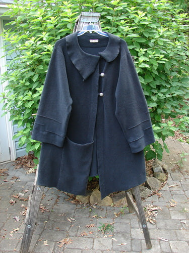 Barclay Celtic Moss Asymmetrical Collar Coat in unpainted black on a clothes rack, showcasing its unique collar, button closure, and elegant varying hemline with belled sleeves from Bluefishfinder.com’s winter collection.