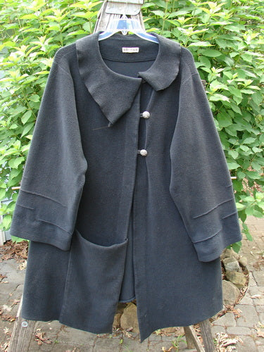 Barclay Celtic Moss Asymmetrical Collar Coat in Unpainted Black, featuring a unique offset collar, three-button closure, varying hemline, belled sleeves, and large exterior pocket, displayed on a swinger.