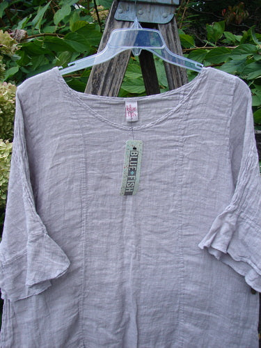 Barclay NWT Gauze Sectional Bias Flounce Top in Rice Paper Pink, featuring a wide scoop neckline, three-quarter sleeves with petal accents, and an A-line shape, displayed outdoors on a hanger.