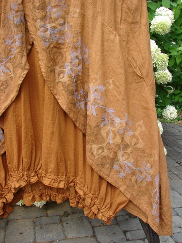 A close-up of a Barclay Linen Open Front Drawcord Hem Jumper in Squash, featuring an interesting layered cross over front painted linen apron with a batiste under skirt layer. The jumper has a downward curved empire waist seam, a deeper oval neckline, and sectional rear panels. The elastic gathered hemline adds a unique touch. Size 2.