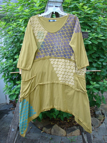 Barclay Raw Seam Vector Tunic Dress in Yellow Olive, featuring a softly rounded V neckline, raw edges, and a varying hemline, displayed outdoors on a clothesline with vibrant patterns.